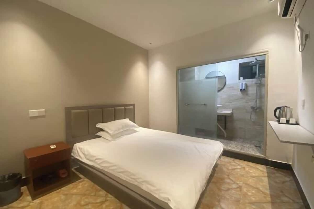A hotel room with a bed, a small wooden nightstand, a mirror, and a view of the bathroom through a glass door.