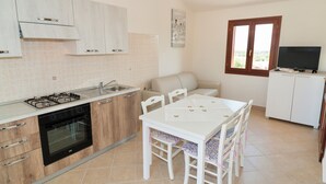 Cocina compacta con estufa a gas, horno y pileta, zona de comedor con mesa y sillas, y un televisor sobre un mueble.