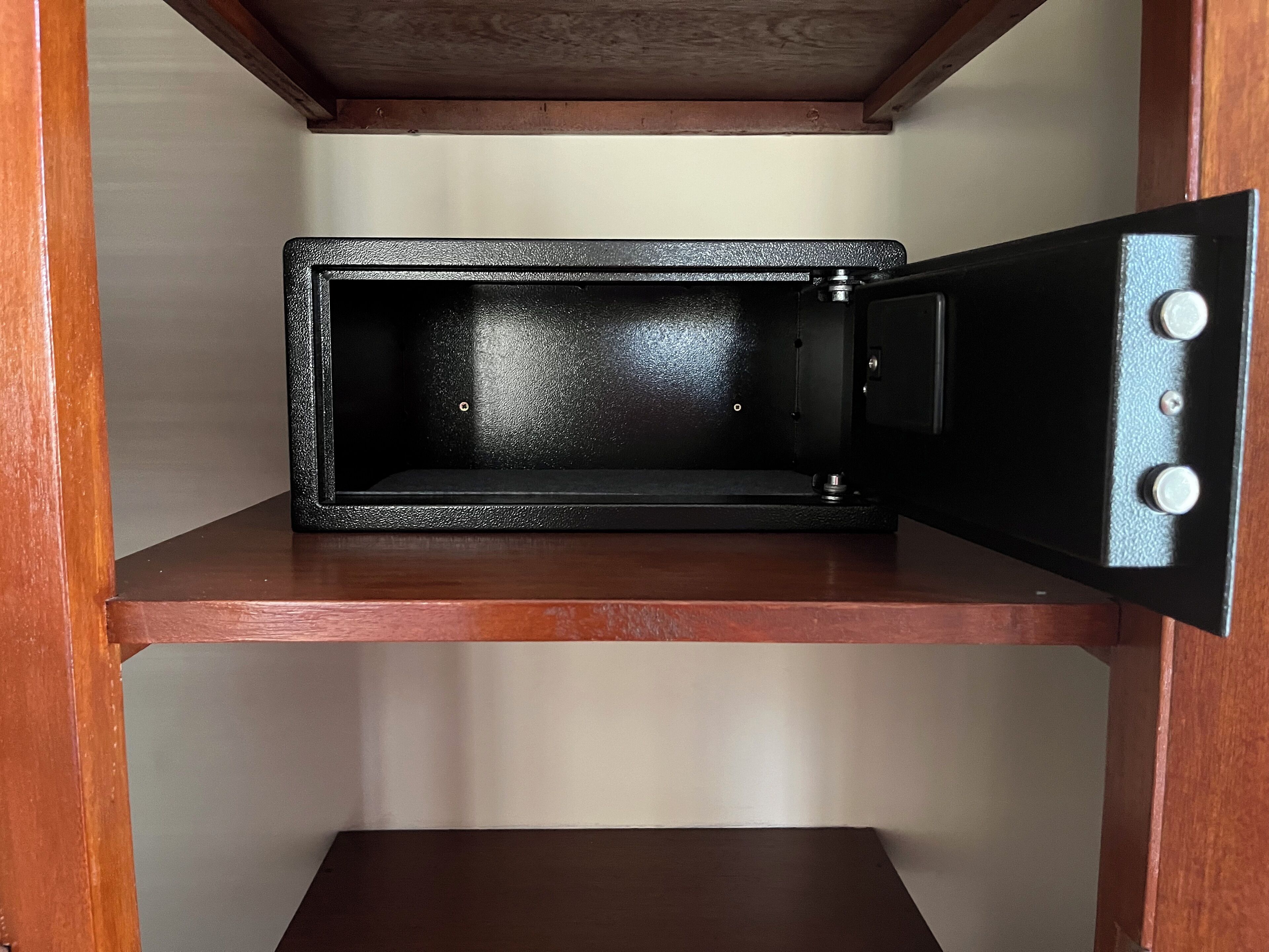 Um cofre preto com puxador metálico e fechadura, colocado sobre uma prateleira de madeira dentro de um quarto.