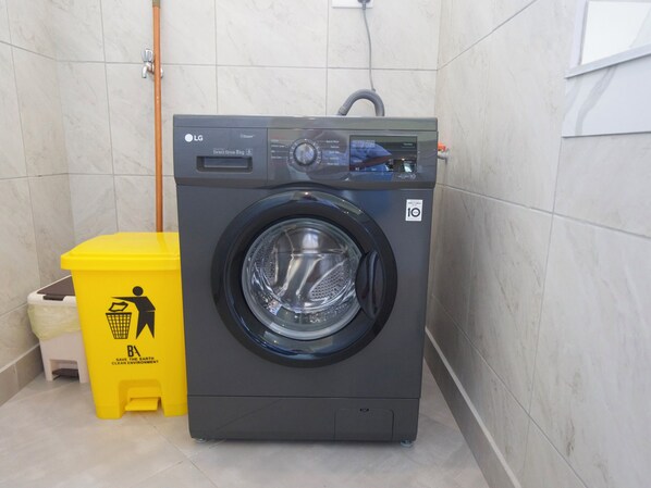 Una lavadora de carga frontal con pantalla digital, un contenedor amarillo para reciclaje y un cubo de basura blanco en una habitación con paredes de cerámica.