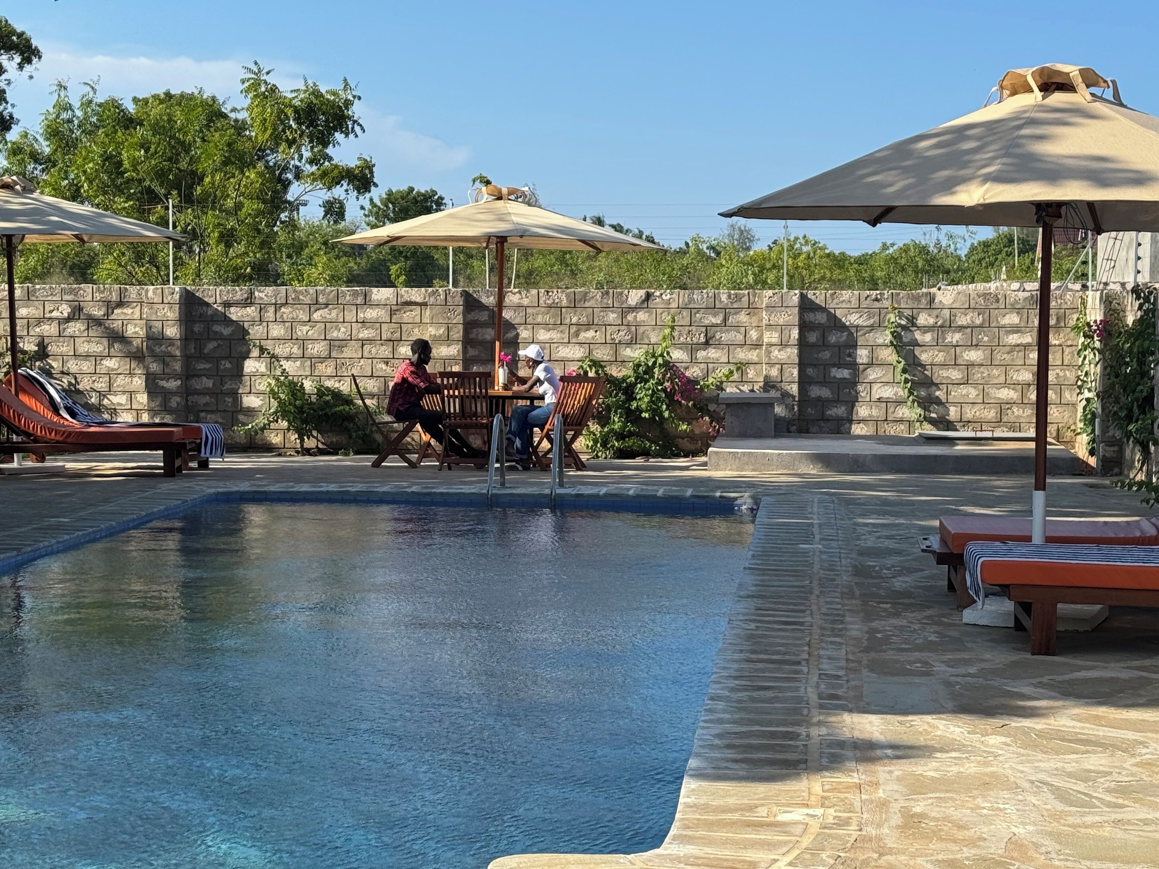Ein Poolbereich mit Liegestühlen und Sonnenschirmen, eine Steinmauer und viel Grün im Hintergrund.