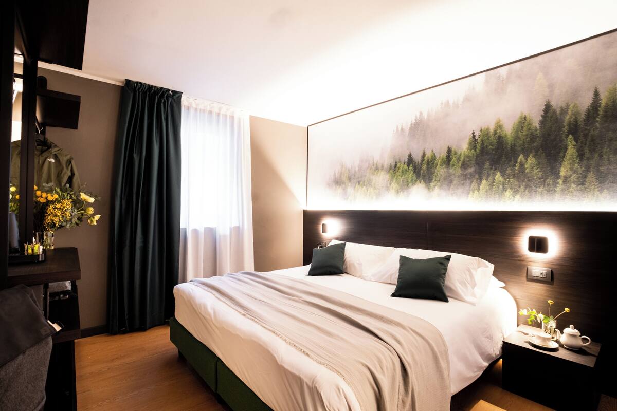 A modern hotel room with a large bed, a nightstand with a lamp, a painting of a forest, and a desk with a chair.