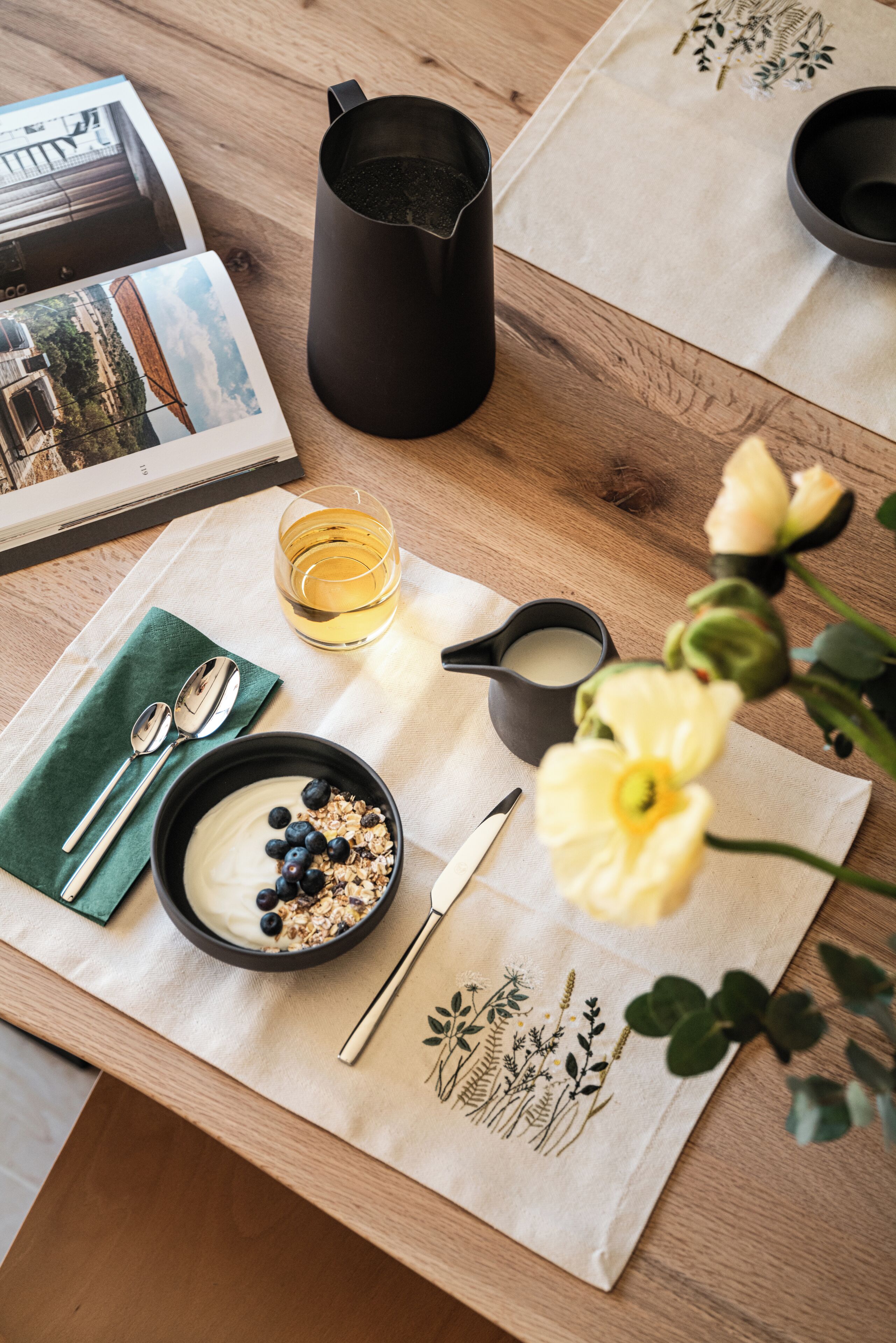 Un desayuno con una mesa de madera, un tazón de yogur con arándanos, un vaso de jugo, una jarra negra, un juego de cucharas y una servilleta decorativa.