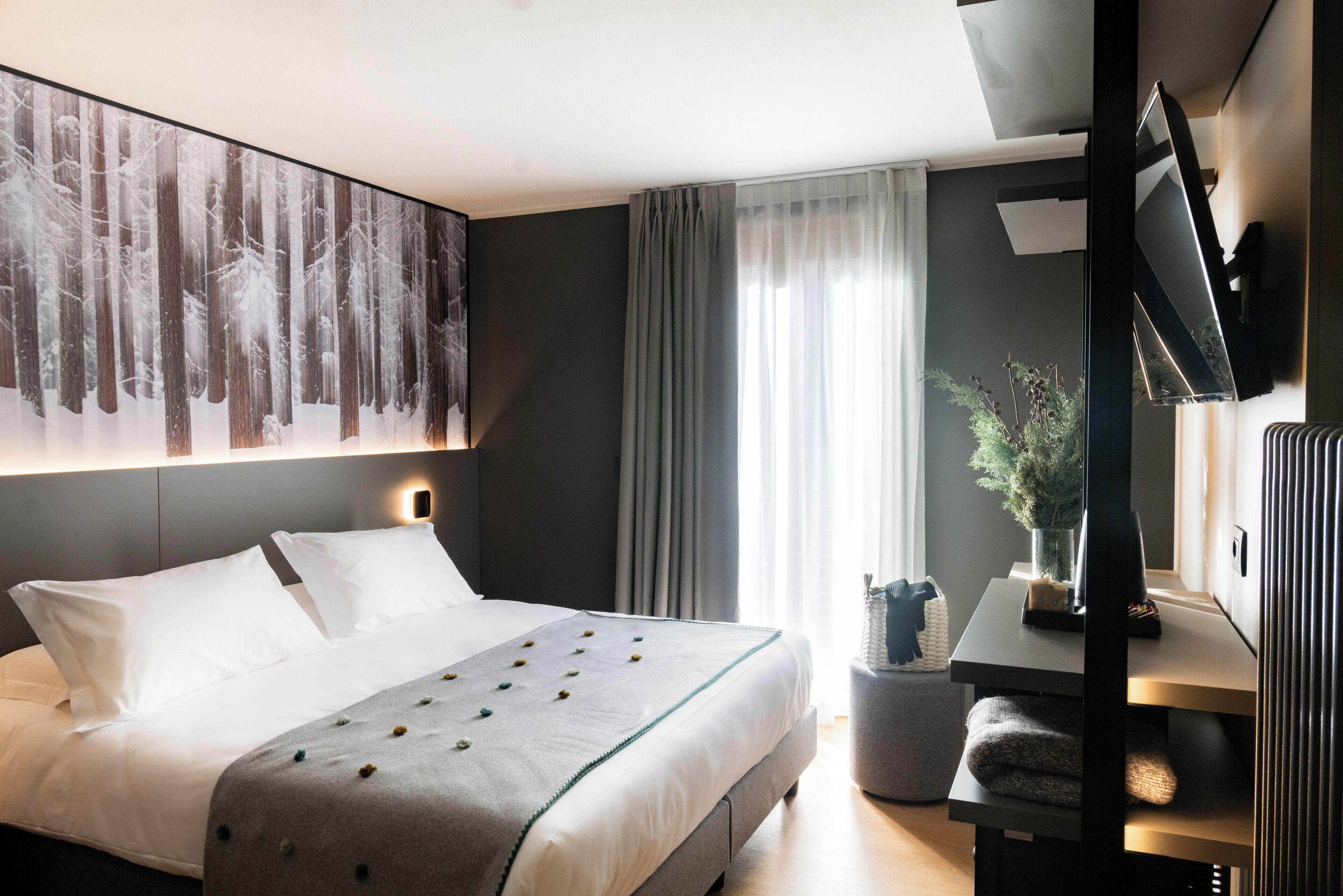 A modern hotel room with a large bed, a mounted TV, a small table with a plant, and a large abstract painting on the wall.