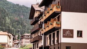 Un hôtel doté de nombreux balcons ornés de plantes, une enseigne indiquant « QUE CIME HOTEL », et un paysage montagneux en arrière-plan.