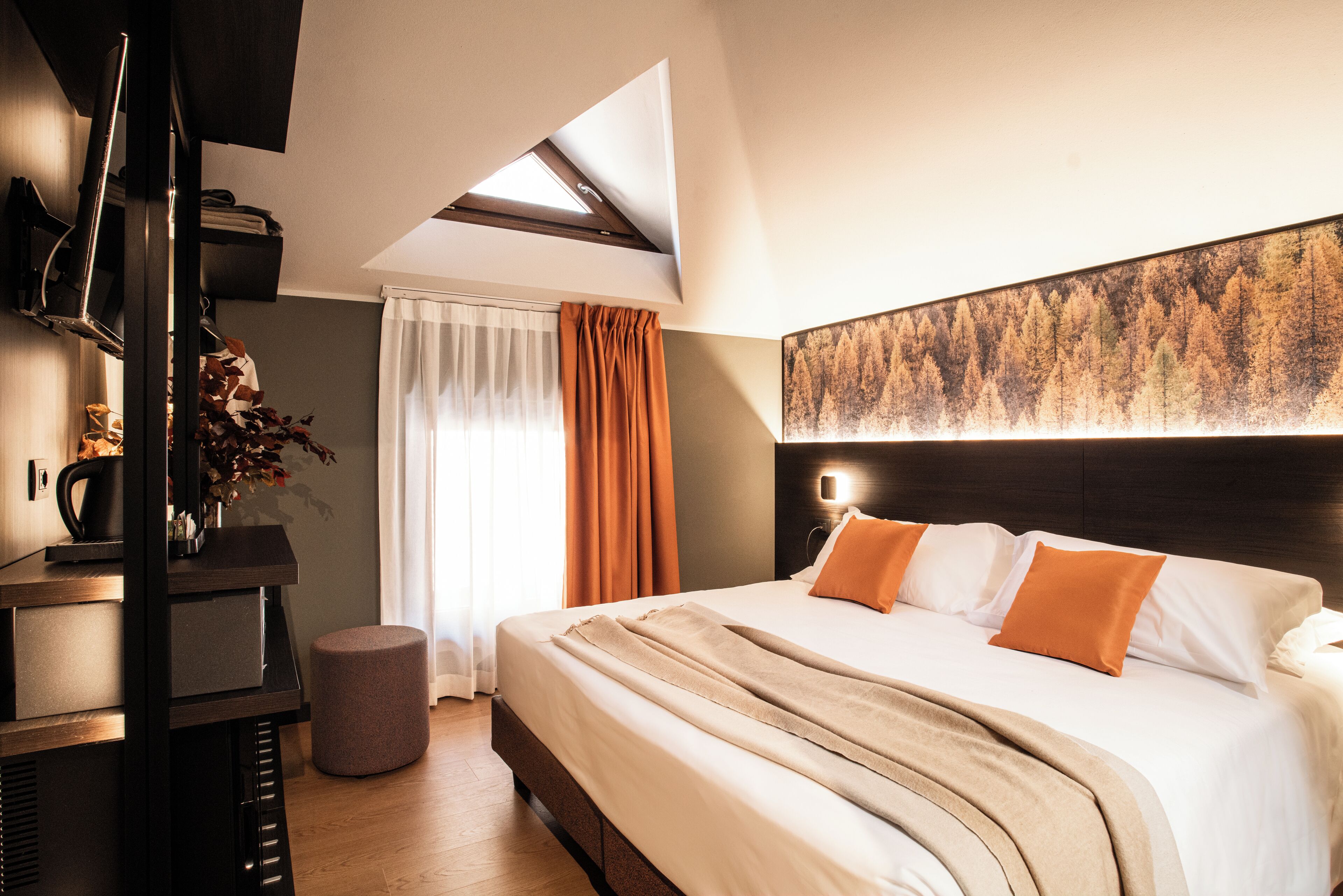 Habitación de hotel moderna con una cama grande, un televisor de pantalla plana, un otoman redondo pequeño y un cuadro en la pared.