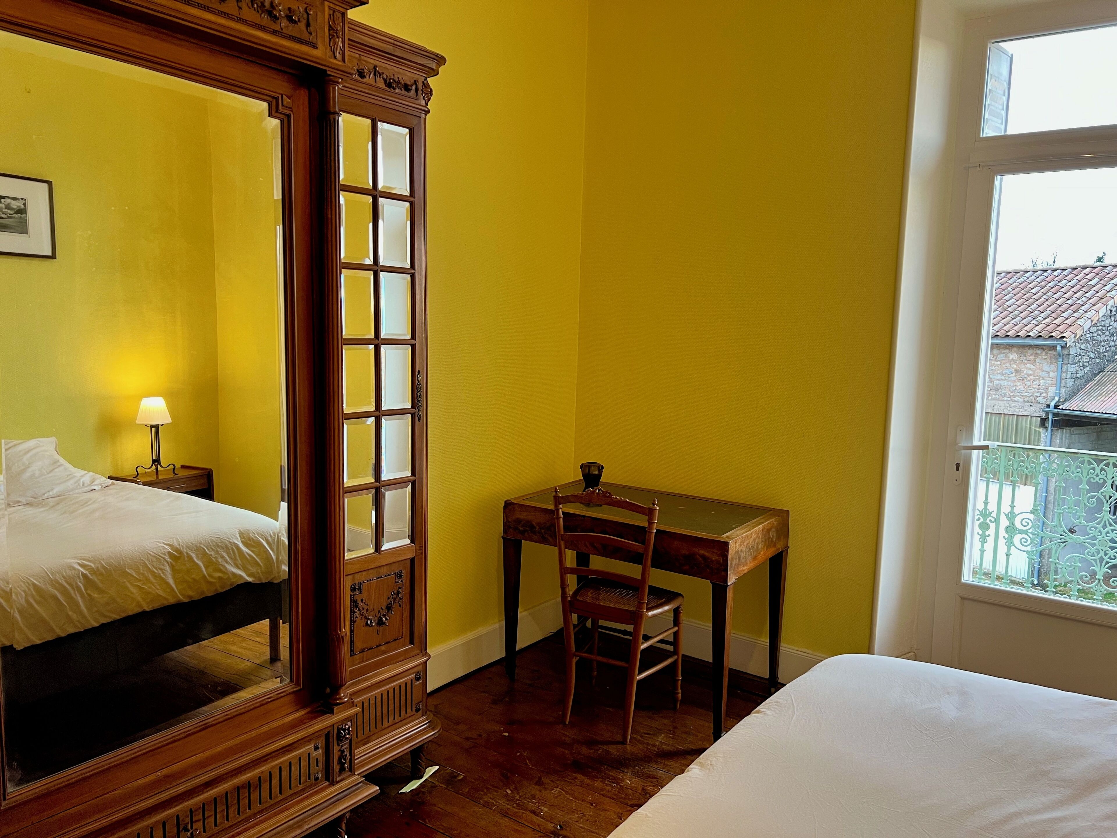 A room with a wooden four-poster bed, a desk with a chair, and a window with a view of buildings.