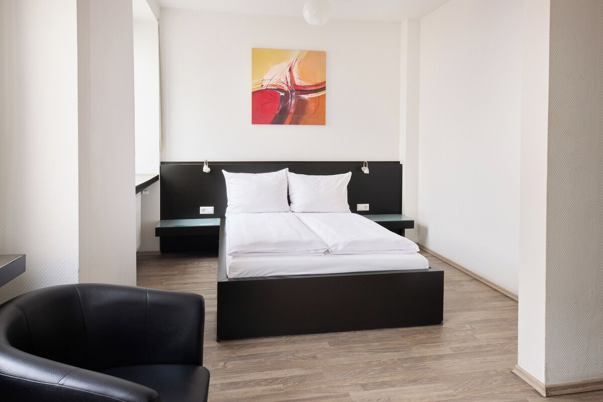 A modern hotel room with a black headboard, white bedding, a single bed, a black leather chair, and a wooden floor.