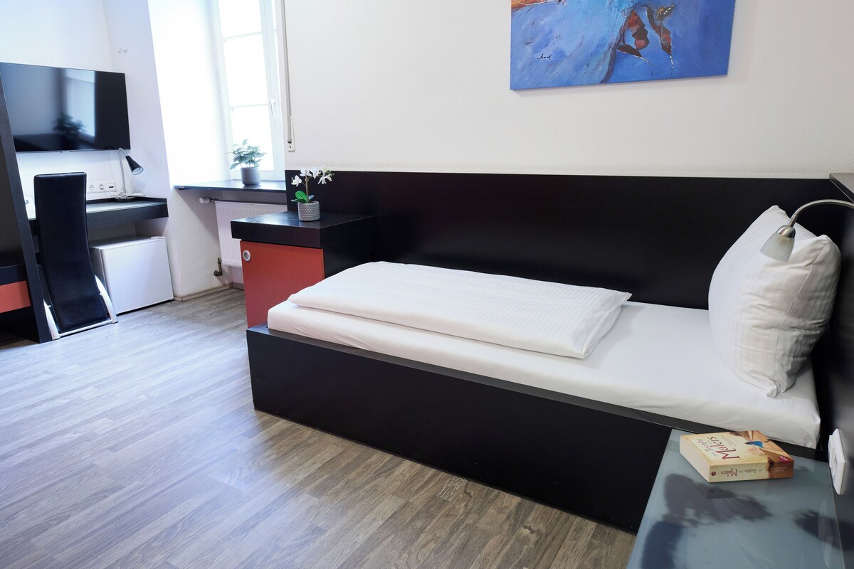 A modern room with a black sofa bed, a small desk with a computer, a potted plant, and a painting on the wall.
