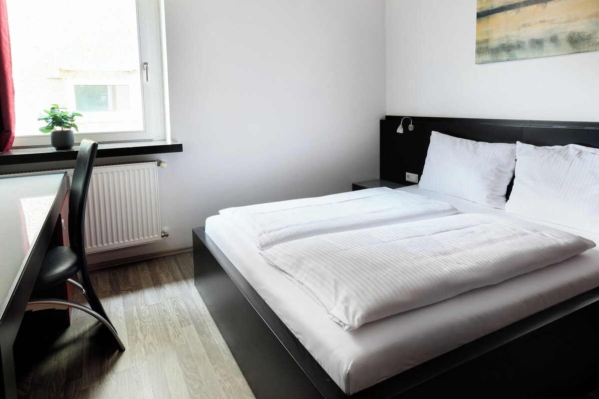 A neatly made bed with white linens, a black headboard, and a potted plant on the windowsill.