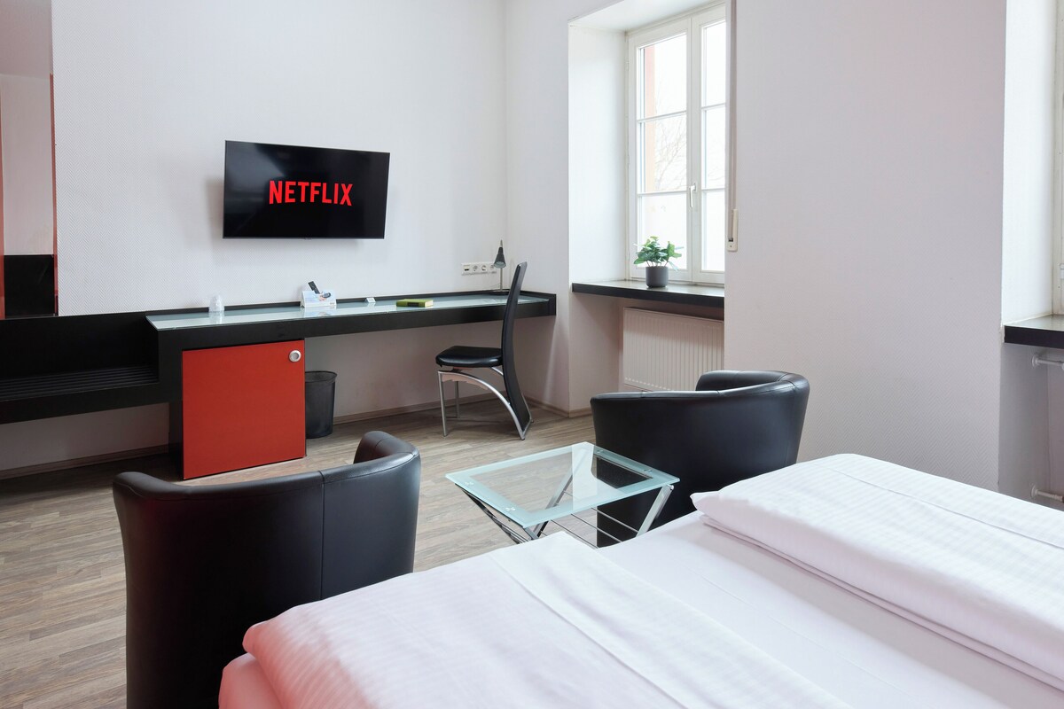 A modern hotel room with a bed, a desk with a computer, a chair, a glass table, and a TV displaying 'NETFLIX'.