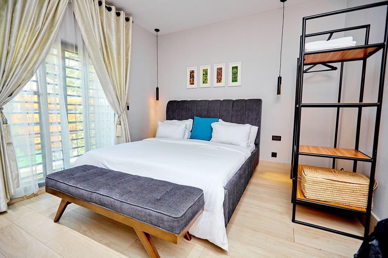Un dormitorio moderno con una cama grande, un banco, un estante en la pared y una ventana con cortinas.