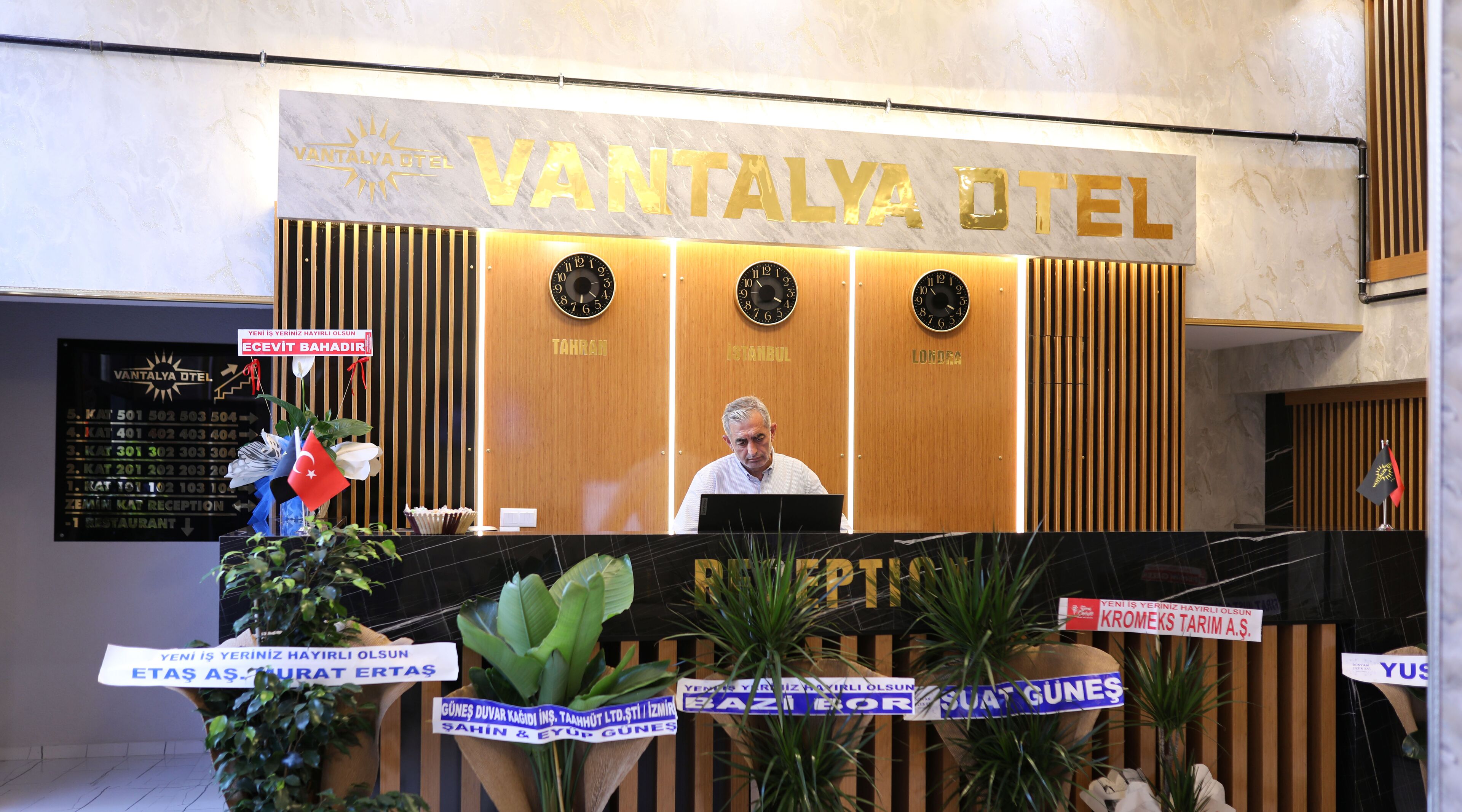 Ein Empfangsschalter im VANTALYA OTEL mit Holzpaneelen, drei Uhren und einem Schild mit Zimmerbezeichnungen.