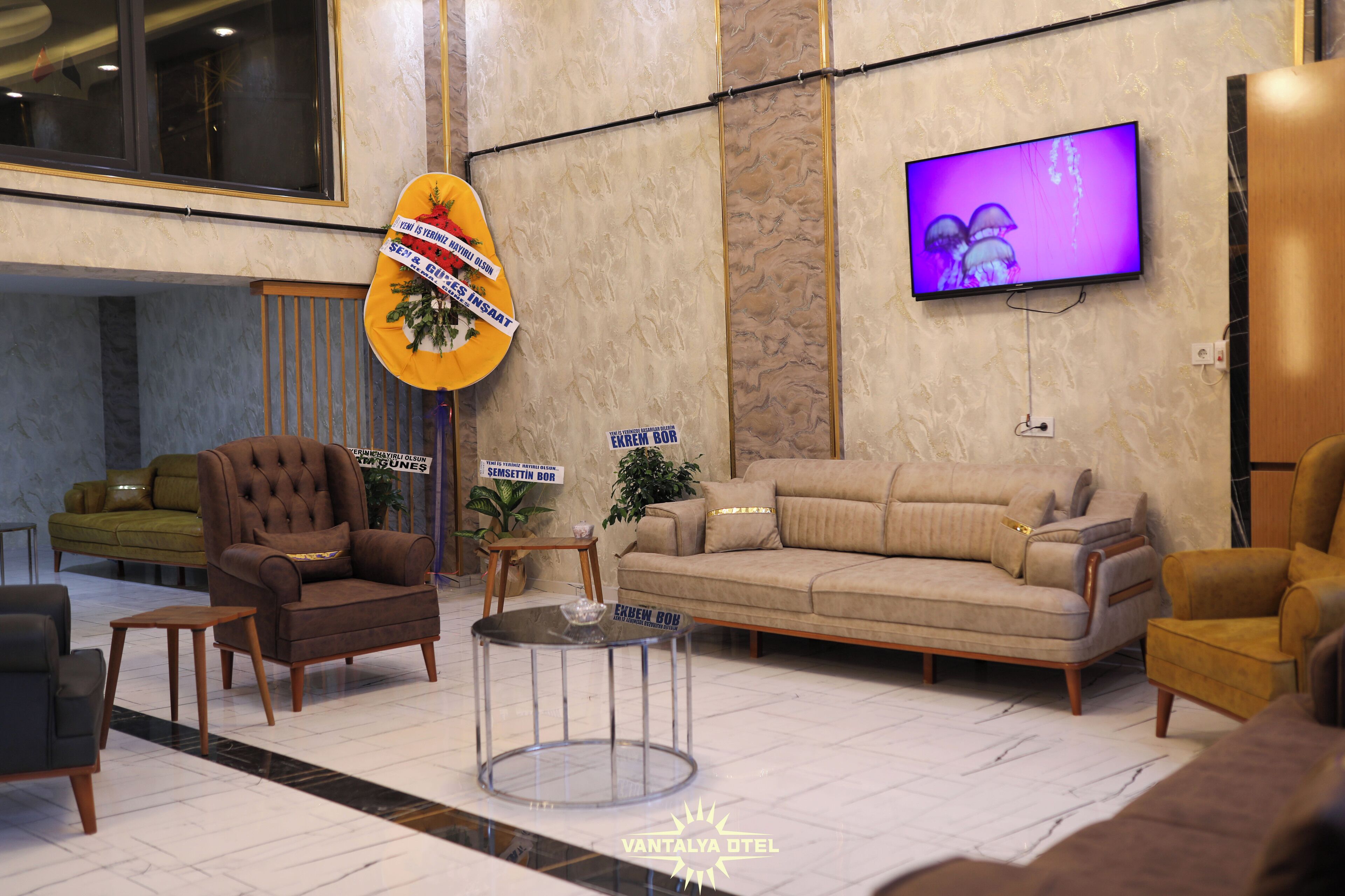 A lobby with a beige sofa, a brown armchair, a small round table, and a television mounted on the wall.