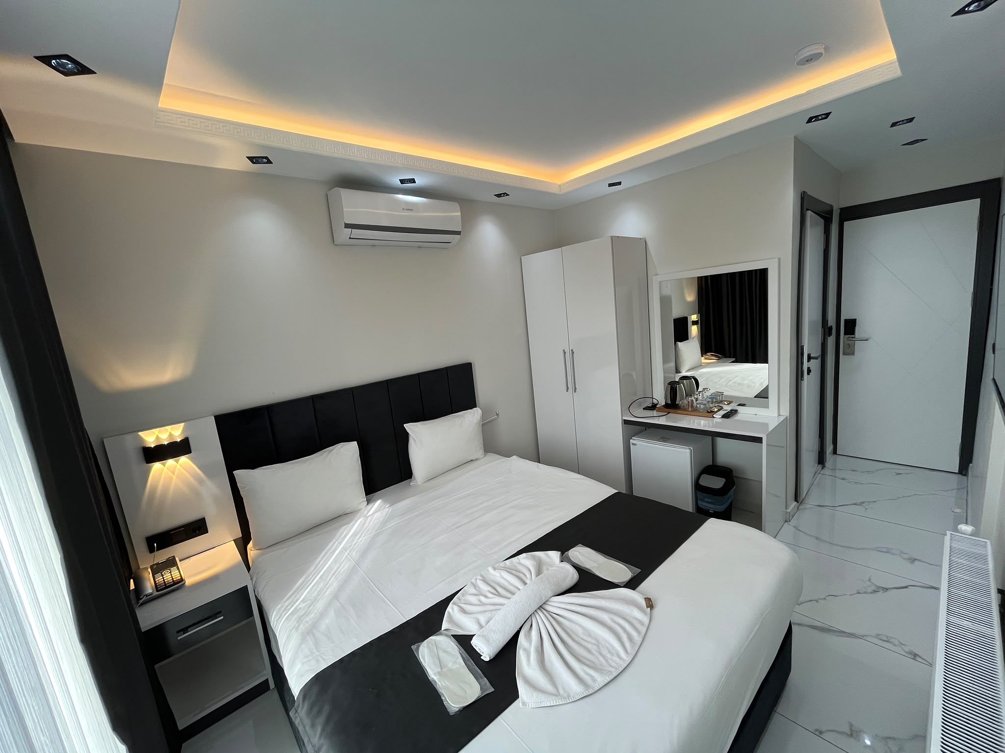 A modern hotel room with a bed, bedside table, air conditioning unit, wardrobe, and a bathroom visible through an open door.