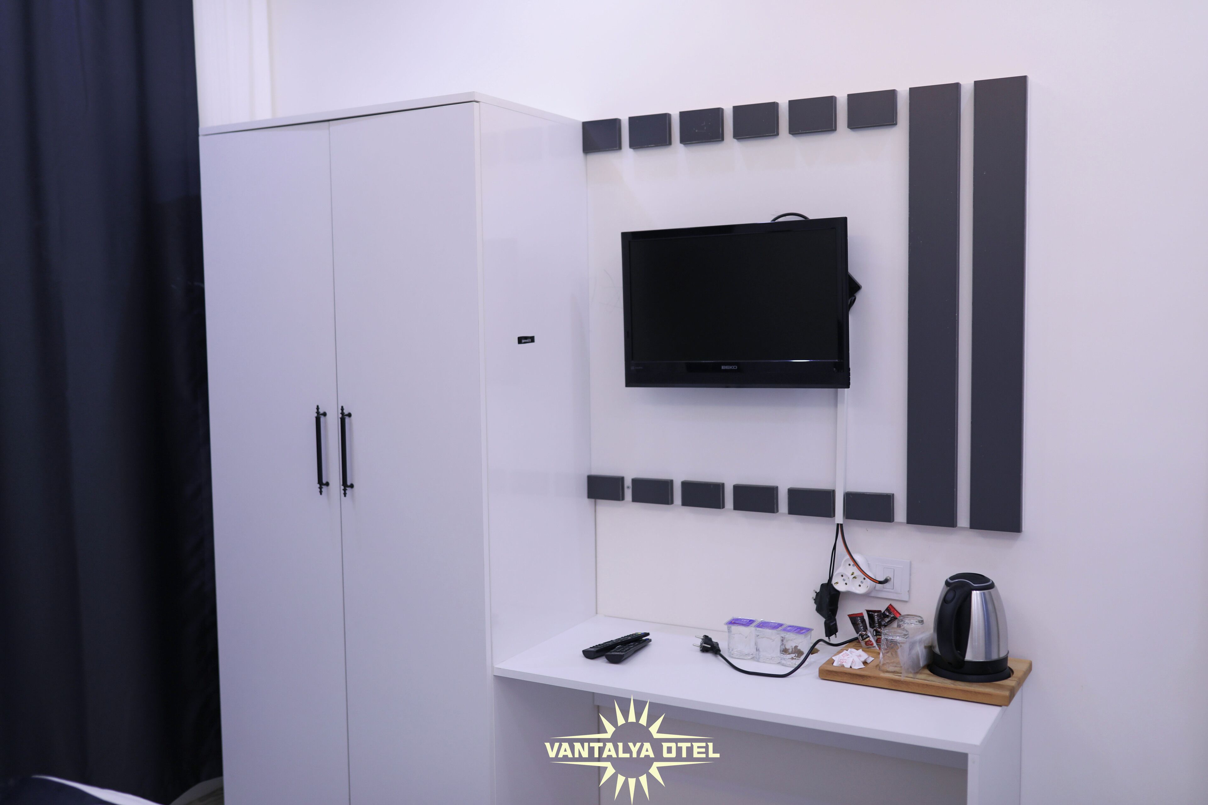 A hotel room with a white wardrobe, a mounted TV, and a small table with a kettle and remote control.
