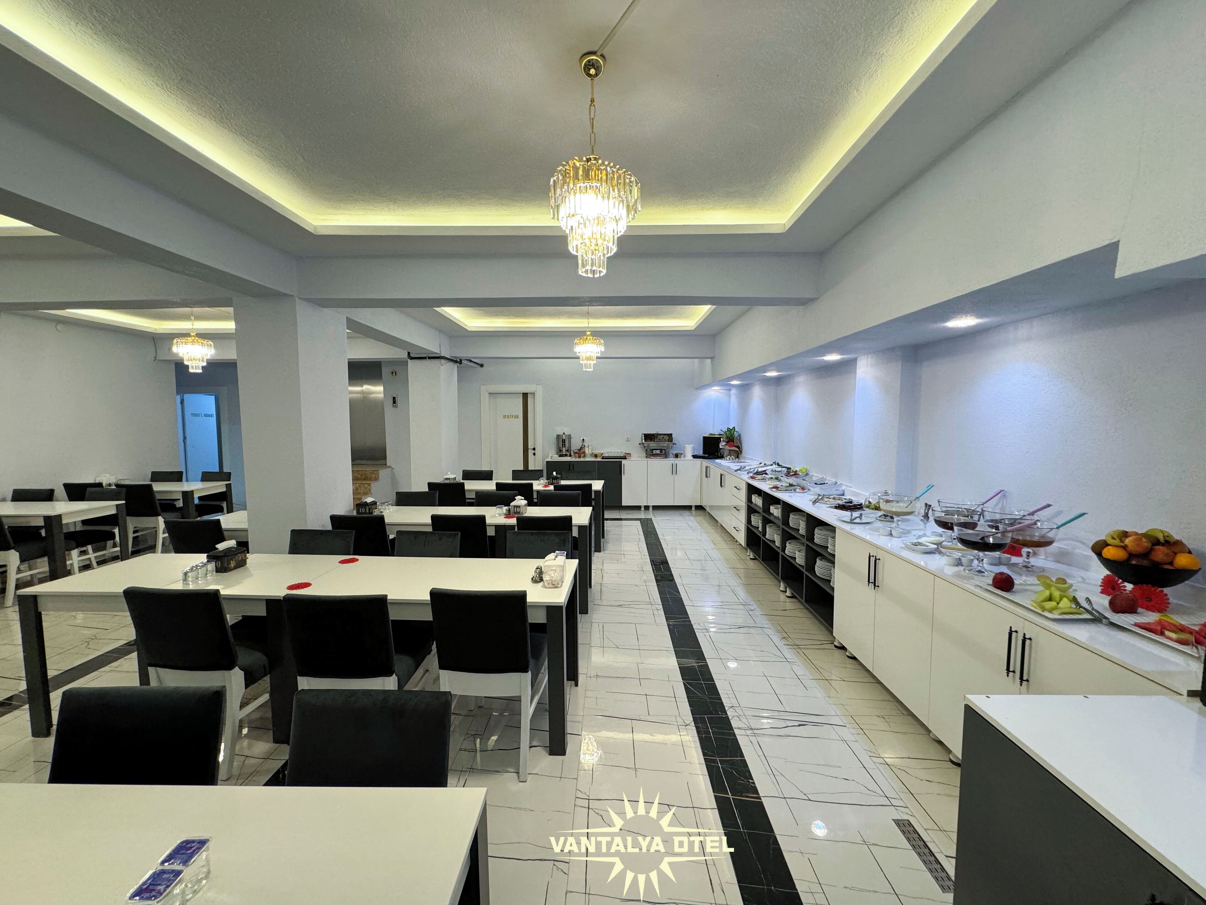 A modern hotel dining area with multiple tables and chairs, a buffet counter with various food items, and a chandelier.