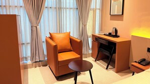 A modern hotel room with a leather armchair, a small round table, a desk with a lamp, and a framed artwork on the wall. - Hotel Fun Regency (Ahmedabad)