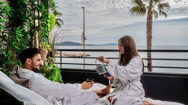 Un couple, vêtu de peignoirs blancs, est assis sur un lit, avec vue sur la mer et les palmiers.