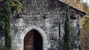 A rustic stone building with a wooden door, a picnic table in front, and surrounding greenery.