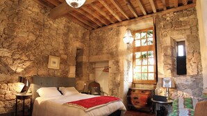 A rustic bedroom with a stone wall, wooden beams, a large bed, a nightstand, a chair, and a window with a view.