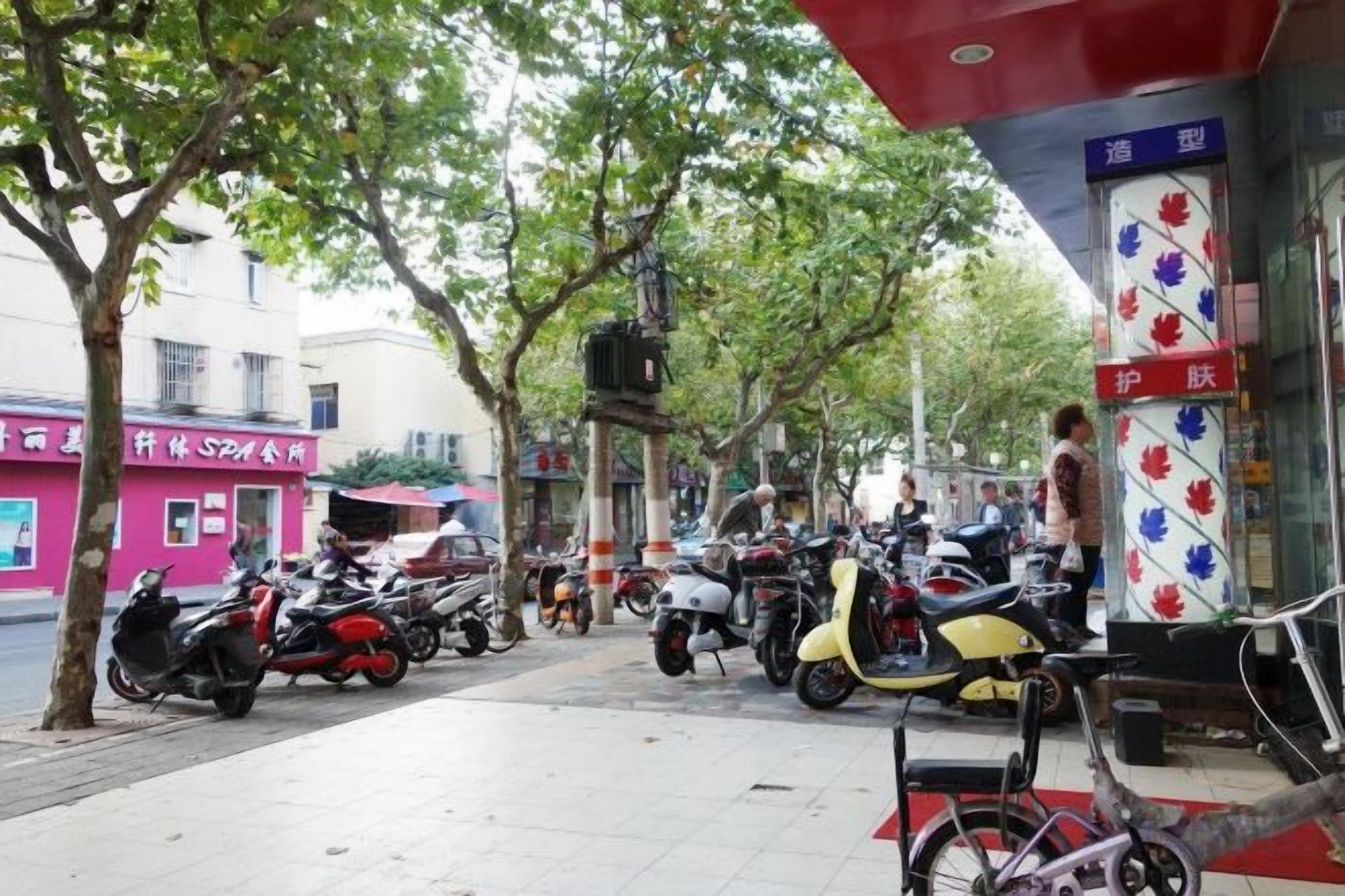 A sidewalk with parked scooters and bicycles, a building with a red and white sign, and trees lining the street.