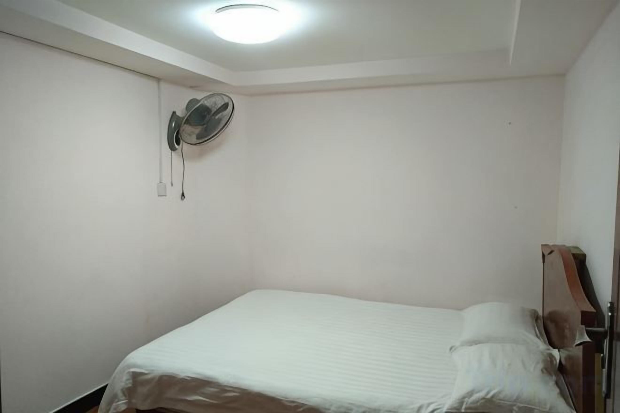 A single bed with a white mattress and pillows, a wall-mounted fan, and a ceiling light fixture.