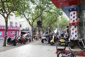A sidewalk with parked scooters and bicycles, a red awning, and a building with a red and white sign. - Huike Hostel (Shanghai)