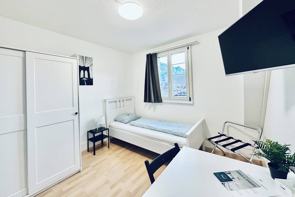 Una habitación pequeña y luminosa con cama, ventana con cortinas, escritorio con silla y una planta.