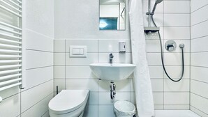 A bathroom with a toilet, sink, shower, and a small trash bin. - ZIMMERzuVERMIETEN.ch Balsthal (Balsthal)