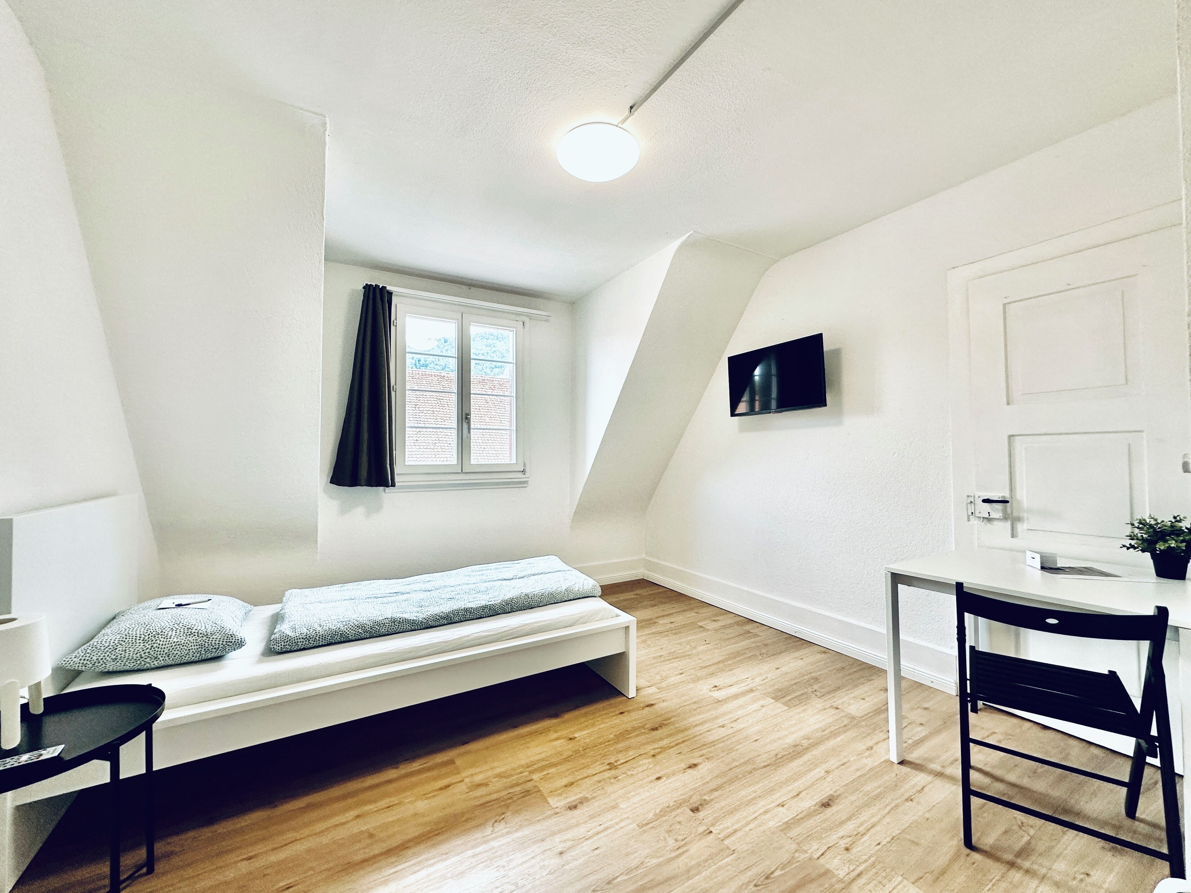 A small, well-lit room with a sloped ceiling, a single window with curtains, a bed, a desk with a chair, and a mounted TV.