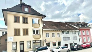 A row of multi-story buildings with various architectural styles, including a beige building with a balcony and a red building with a sloped roof. Several cars are parked along the street in front of the buildings. - ZIMMERzuVERMIETEN.ch Balsthal (Balsthal)