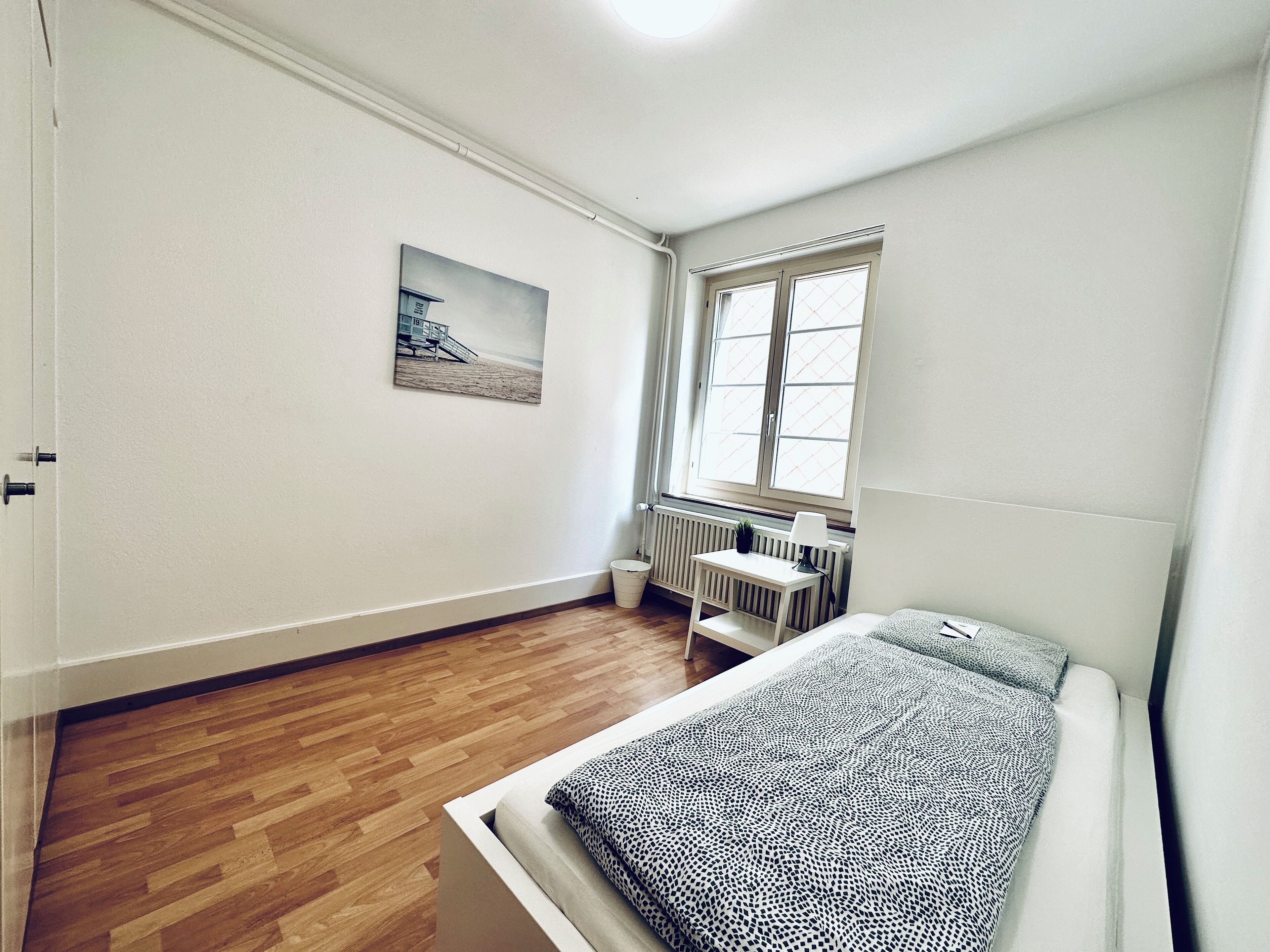Une chambre avec un plancher en bois, un lit recouvert d’une couvre-lit à motifs, un radiateur, une fenêtre avec des stores et un tableau accroché au mur.