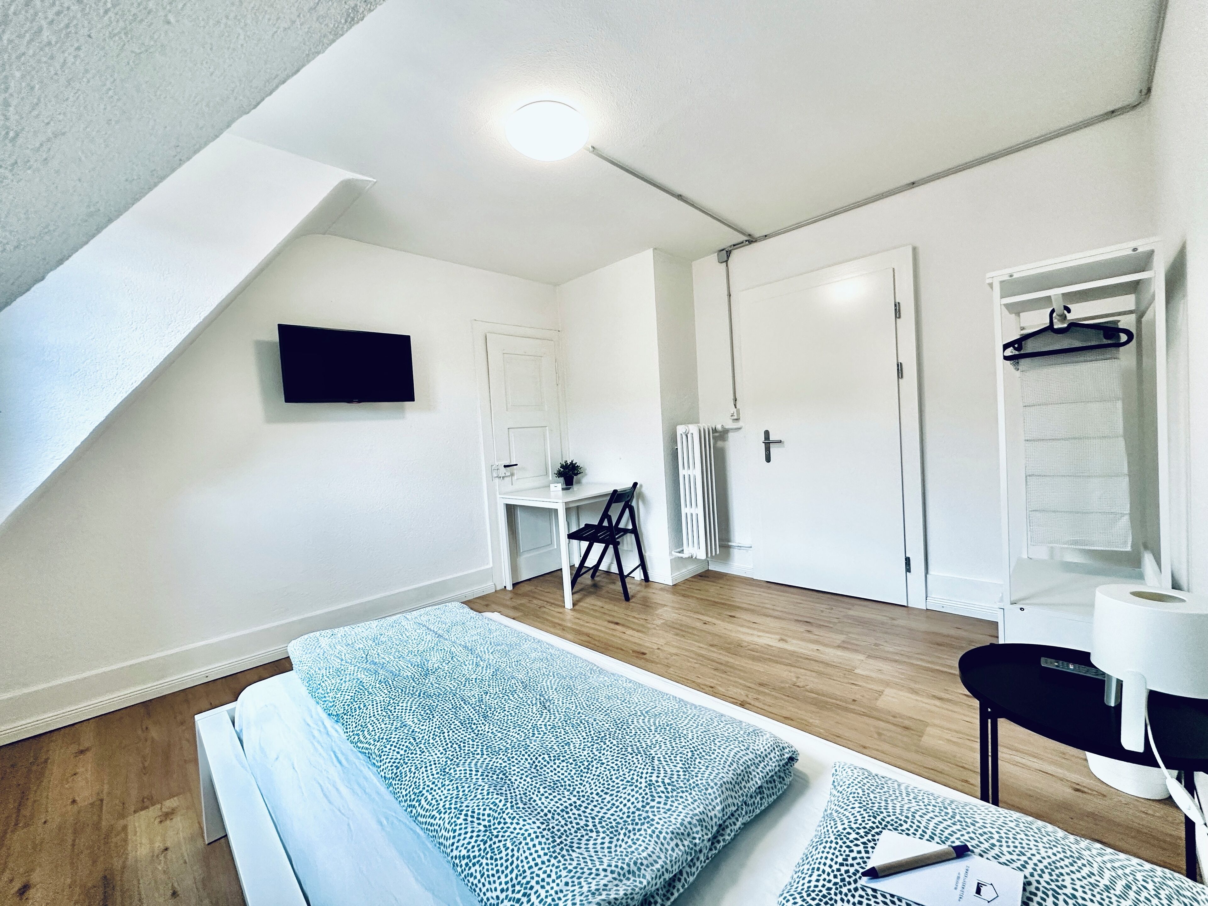 A small, bright room with a bed, a desk, and a chair. There is a TV on the wall, a radiator, and a wardrobe.