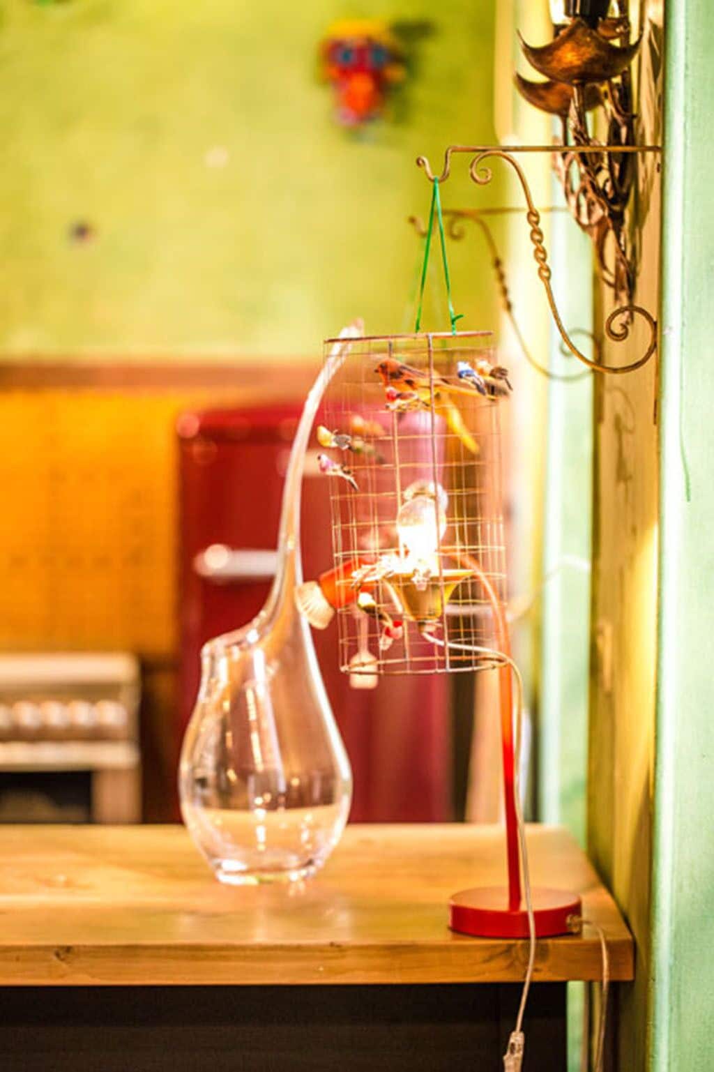 A clear glass vase on a wooden table with a birdcage hanging from the wall.