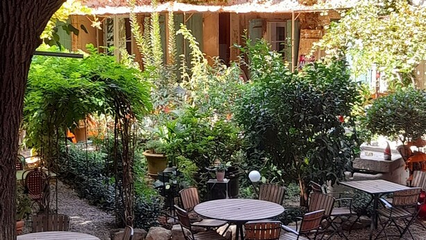 Un área de estar al aire libre con mesas y sillas, rodeada de follaje y un edificio con paredes cubiertas de hiedra.
