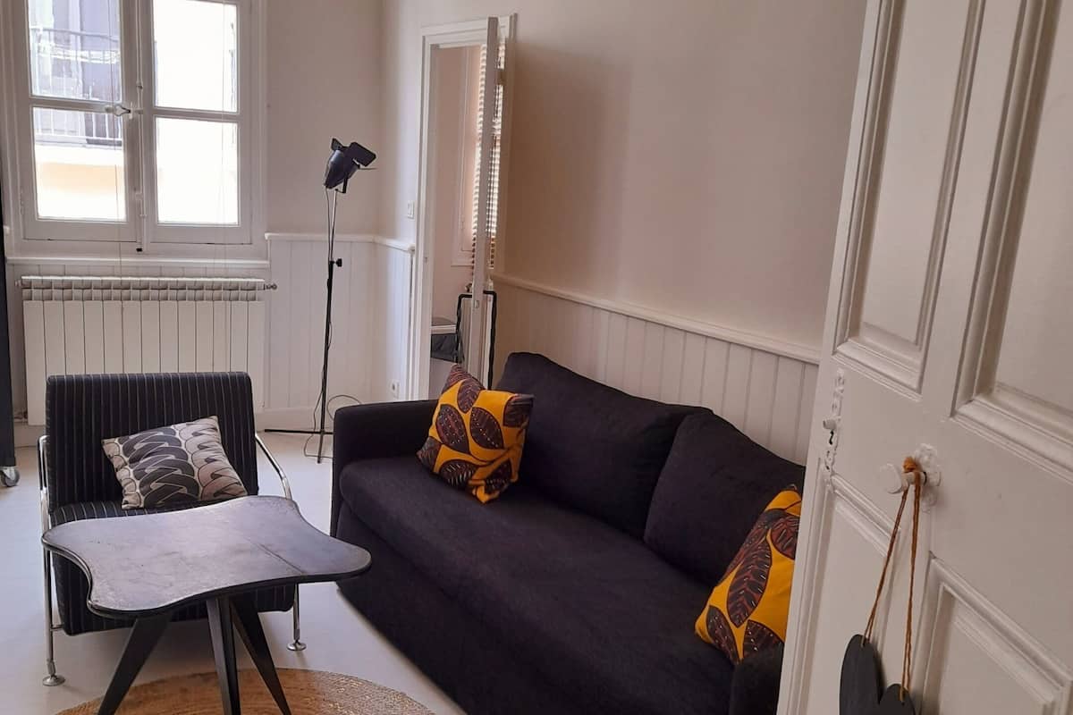 A living room with a black sofa, striped armchair, and a small table. There is a radiator, a standing lamp, and a window with blinds.