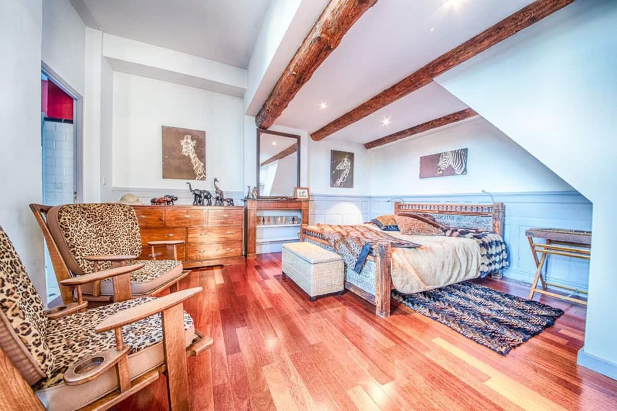 A bedroom with a wooden ceiling, a bed, a chair, a dresser, and artwork on the walls.