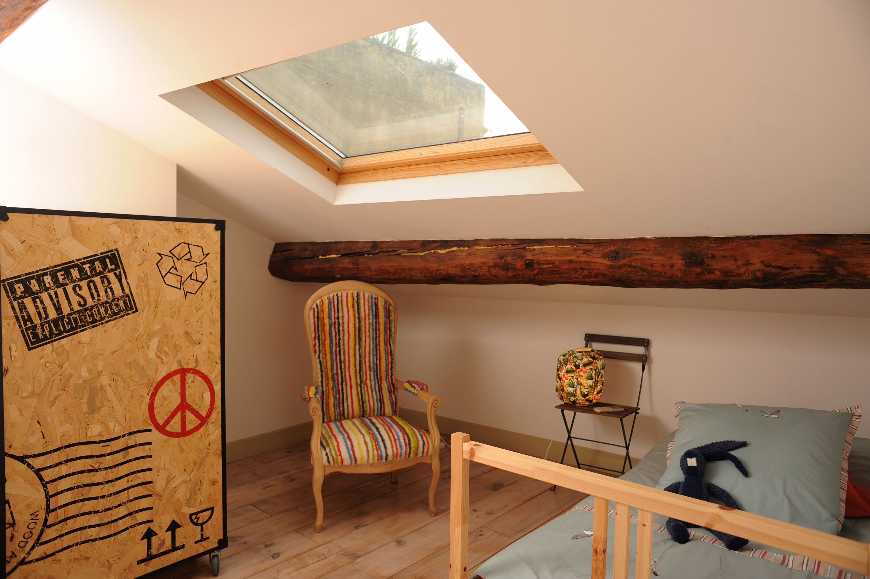 A room with a skylight, a striped chair, a bed with a pillow, and a small table with a floral chair.