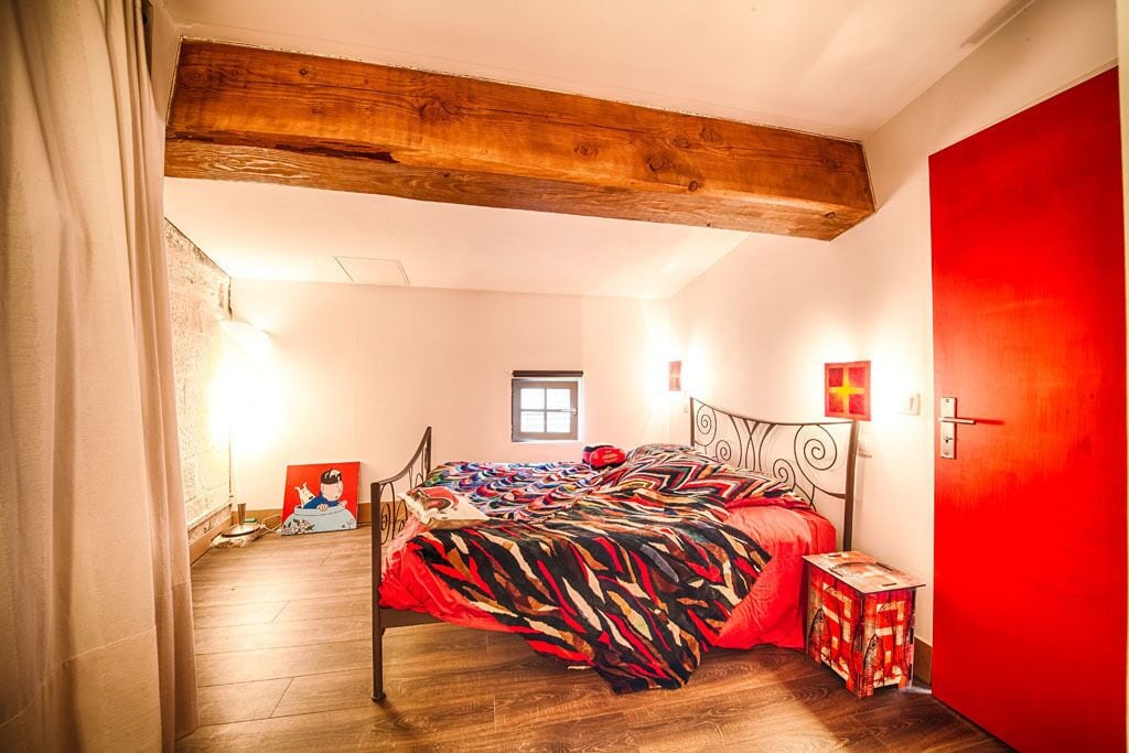 Ein Schlafzimmer mit Holzboden, einer roten Tür, einem Bett mit bunter Decke und einem kleinen Tisch mit einer roten Schachtel.