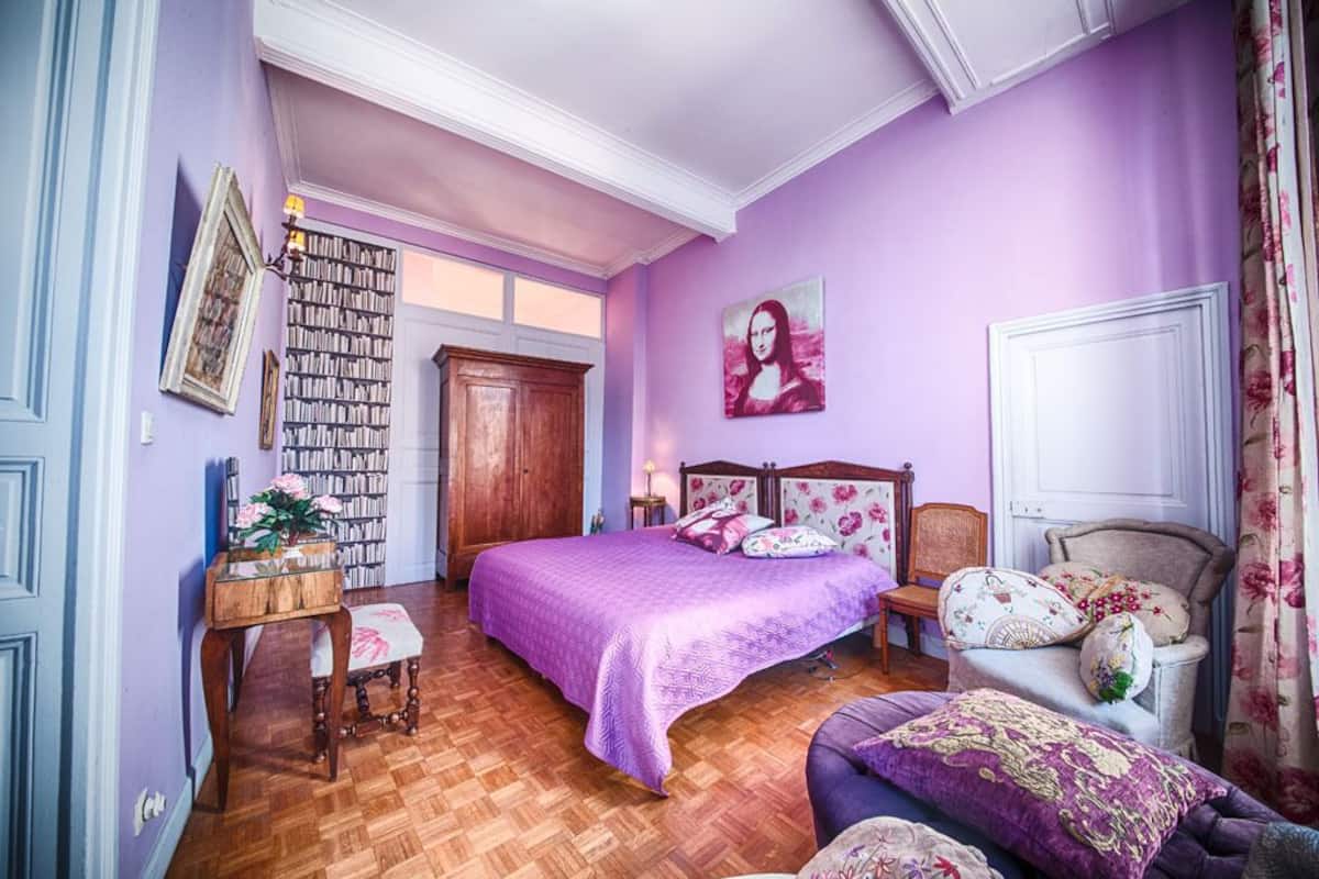 A bedroom with a large bed, a wooden wardrobe, a desk with a chair, and a bookshelf.