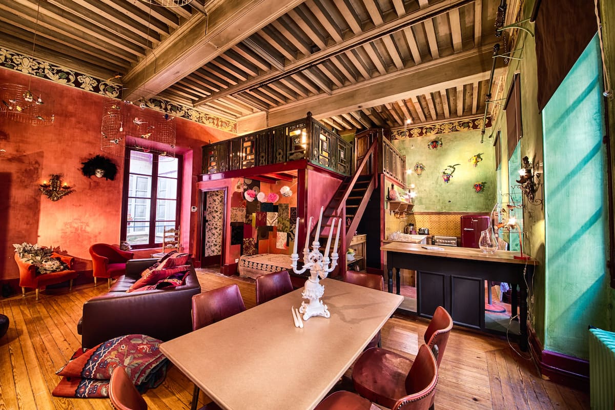 A spacious room with a high ceiling, wooden beams, a dining area with a table and chairs, a staircase, and a kitchen area.