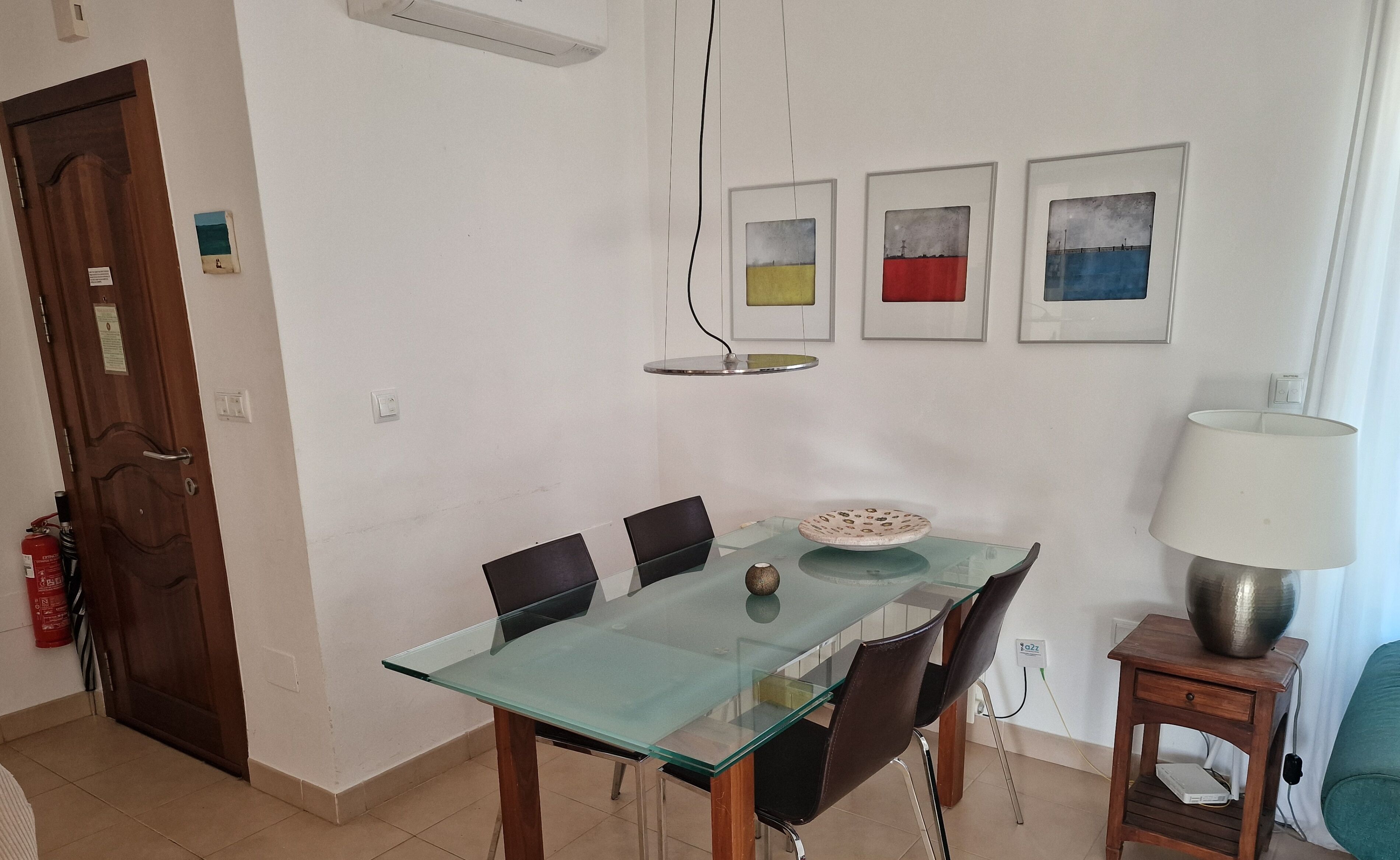 A dining area with a glass table, black chairs, and a small side table with a lamp. There are three framed artworks on the wall and a door with a fire extinguisher.