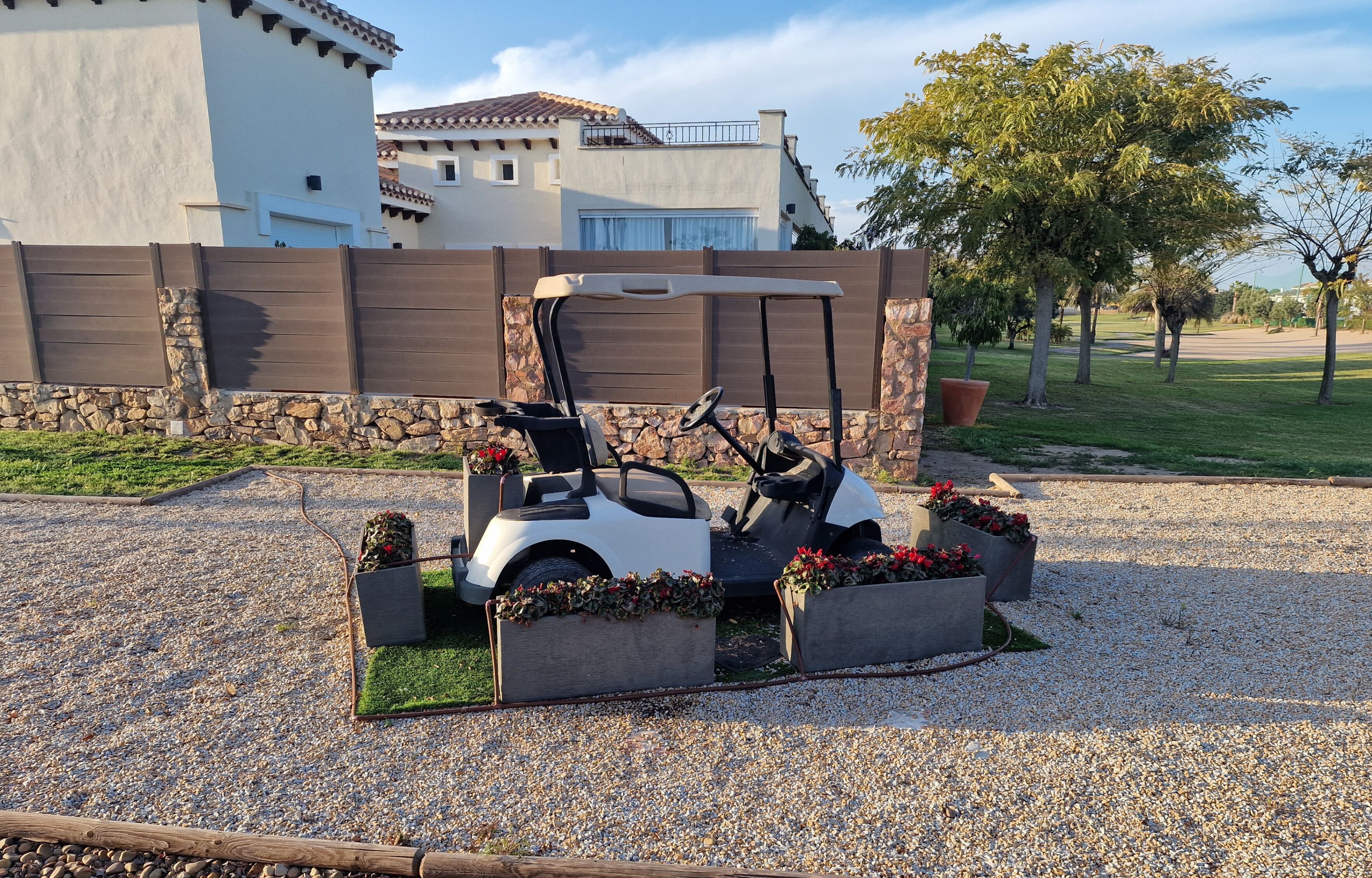A golf cart parked in a gravel area with flower planters, a stone wall, and a building with a tiled roof in the background.