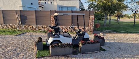A golf cart parked in a gravel area with flower planters, a stone wall, and a building with a tiled roof in the background.
