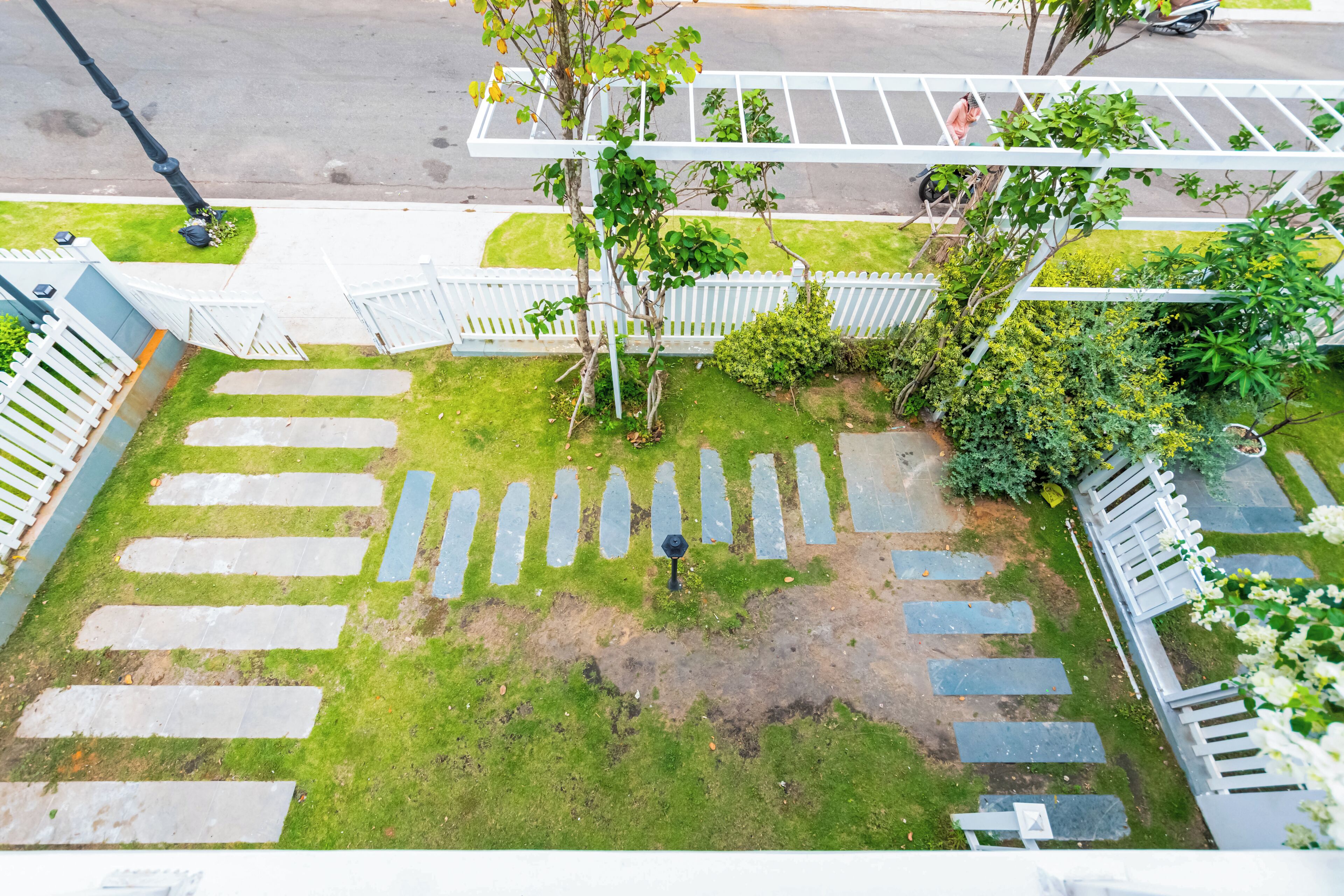 A courtyard with a crosswalk leading to a gated entrance, surrounded by greenery and a white fence.