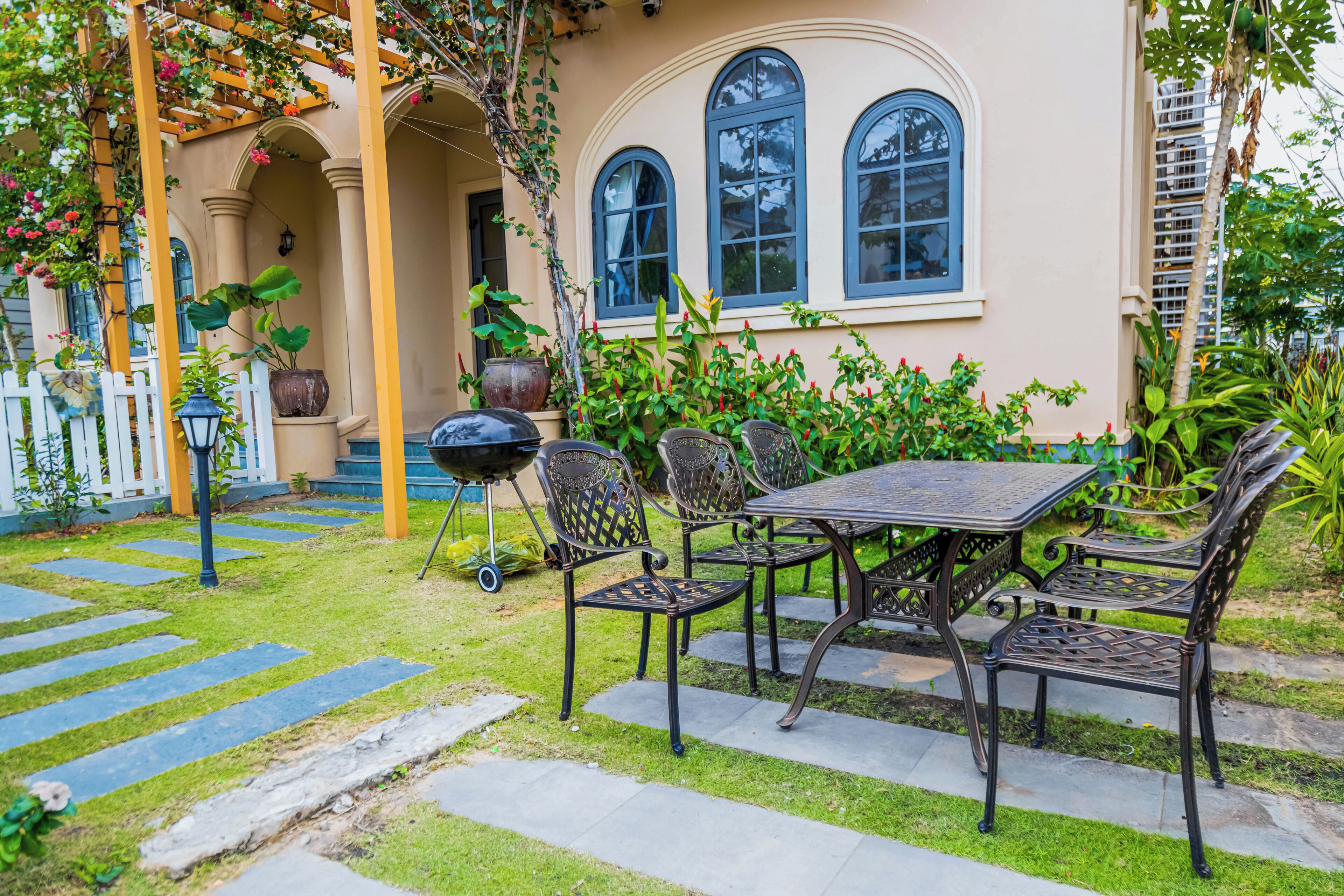 A patio with a table and chairs, a pergola with climbing plants, and a white picket fence.