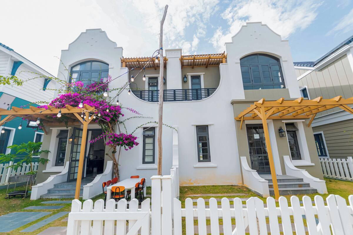 Una casa de dos pisos con fachada blanca, un porche con pérgola de madera y una cerca de piquetes blancos.