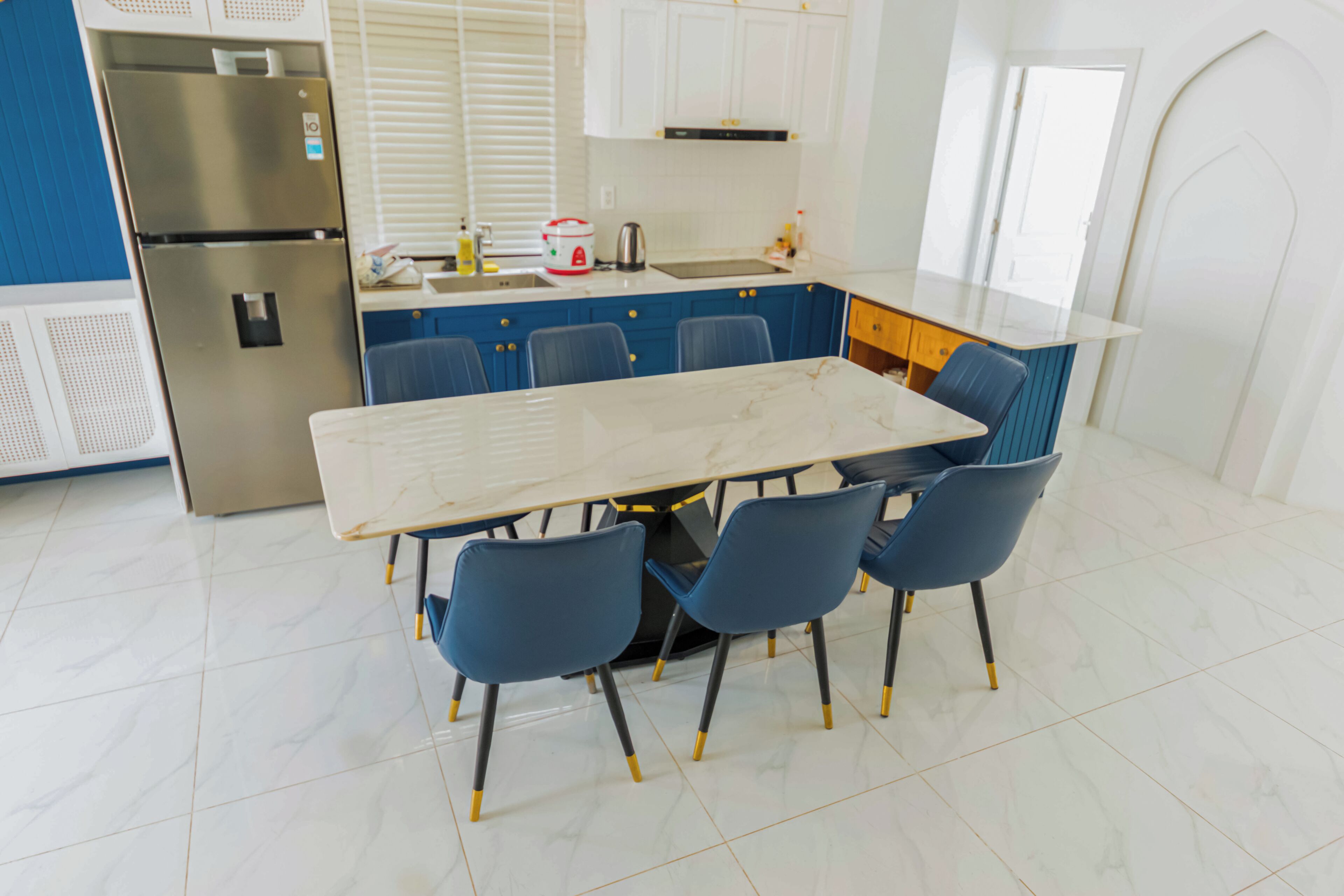 Una cocina moderna con encimera de mármol, refrigerador de acero inoxidable, sillas azules y armarios blancos.