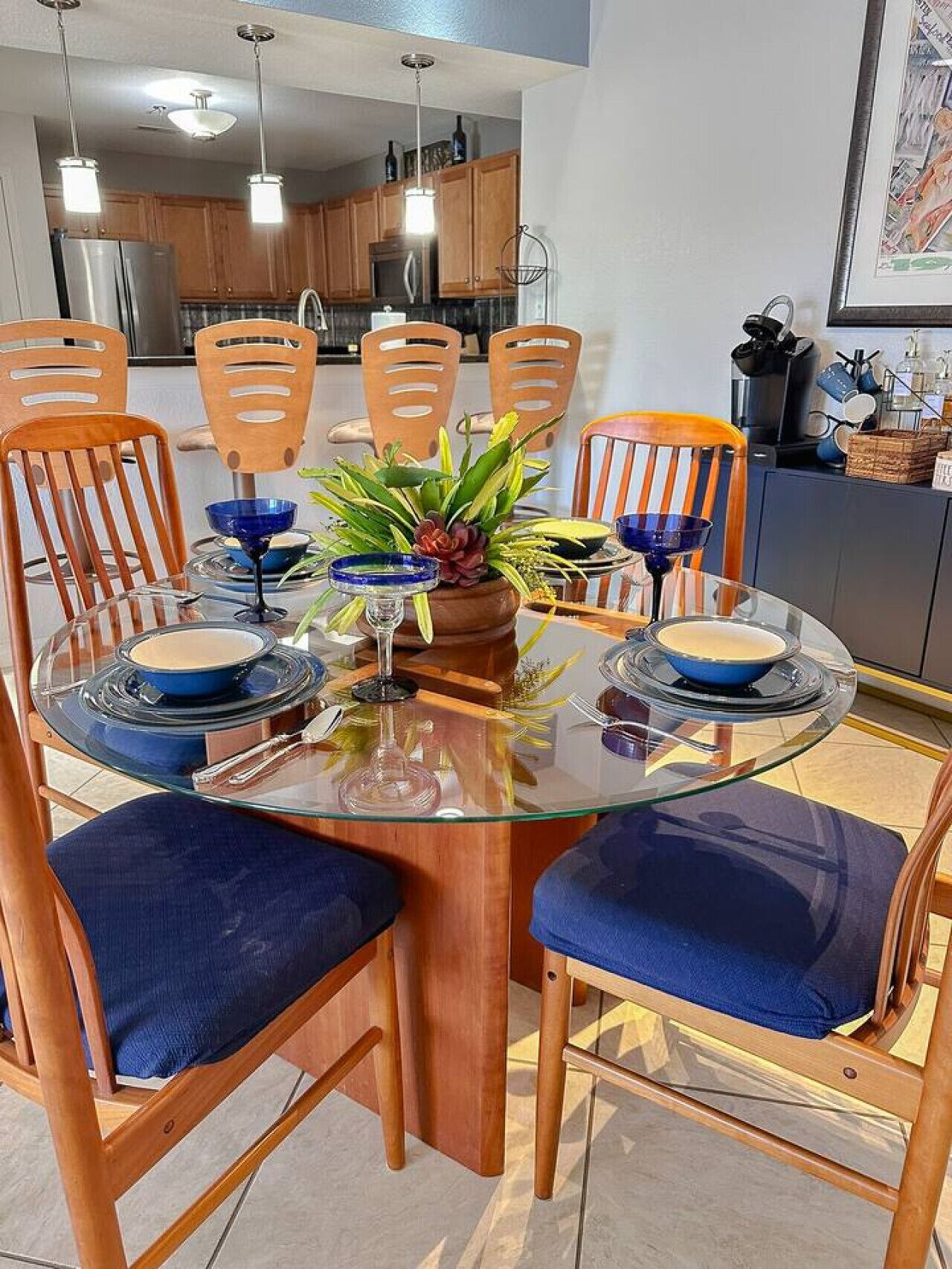 A dining area with a glass table surrounded by wooden chairs with blue cushions, a central plant, and blue dishes.